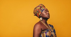 Ghanaian woman wearing traditional clothing against a yellow background. Mature black woman dressed in colourful Kente cloth and golden jewellery. Woman embracing her rich West African culture.