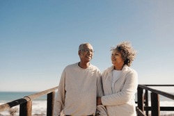 Mature couple smiling happily while taking a walk along a wooden foot bridge at the beach. Romantic elderly couple enjoying a refreshing seaside holiday after retirement.