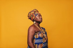 Mature woman wearing an African traditional attire against a yellow background. Confident black woman dressed in Kente clothing and golden jewellery. Woman embracing her rich West African culture.