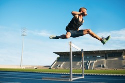 Professional male track and field athlete during obstacle race. Young athlete jumping over a hurdle during training on racetrack in athletics stadium.