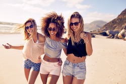 Portrait of three young female friends walking on the sea shore looking at camera laughing. Multiracial young women strolling along a beach.