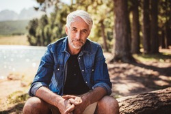 Portrait of mature man sitting near a lake staring at camera. Senior caucasian man relaxing on a log by the lake on a summer day.
