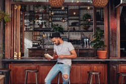 Portrait of trendy young man sitting at a cafe counter reading a book and drinking coffee