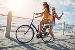 Two stylish young female friends on a bicycle along seaside. Best friends enjoying a day on bike.