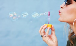 Close up side view shot of young female model blowing soap bubbles on blue background. Focus on hands and wand.