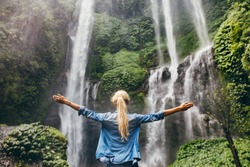 Rear view of young woman standing in front of waterfall with her hands raised. Female tourist with her arms outstretched looking at waterfall.