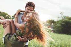 Shot of young woman being carried by her boyfriend in grass field. Couple having fun on their summer holiday.