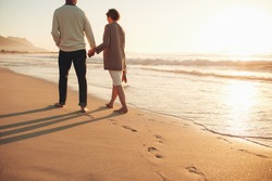 Rear view of a senior couple walking along the sea shore. Mature man and woman together strolling on the sea shore at sunset.