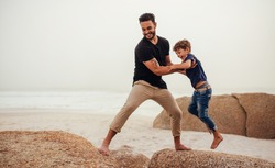 Shot of father helping his son to jump over the rock on the beach. Young man and little boy having fun at the rocky beach.
