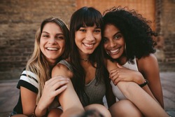 Three beautiful smiling girlfriends taking selfie with mobile phone. Multi ethnic group of women sitting outdoors by the city street and taking self portrait.