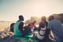 African young man singing and playing guitar on the beach. Group of four people having great time at the beach picnic.