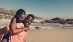 Young african man carrying his girlfriend on his back at the beach. Loving couple having piggyback fun on seaside.