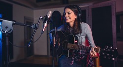 Happy young woman recording a song in a professional music studio. Smiling female sitting on front of microphone with guitar in recording studio.