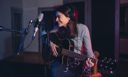 Professional musician recording in studio. Female guitarist sitting in front of microphone and smiling. Singing song for album in recording studio.
