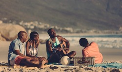 African man playing guitar for friends on the beach. Group of friends having picnic at the seashore.