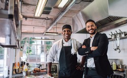 Portrait of restaurant owner with chef in kitchen. Businessman with professional cook standing together and looking at camera.