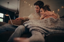 Young couple sitting on sofa at home in winter and using laptop computer. Cozy couple in hygge house with laptop.