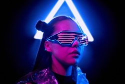 Portrait of serious cyber woman in wire LED glasses standing in light of neon sign