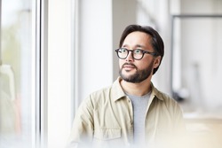 Serious contemplative young Asian man in casual shirt and eyeglasses sitting in office and looking out window