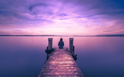 Self reflection in magical world of fantasy. One woman sits on a wooden pier. Cloudy above the lake. Long exposure
