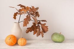 Autumn season concept. Dry oak leaves in vase with decorative orange and green pumpkins on light wooden table.