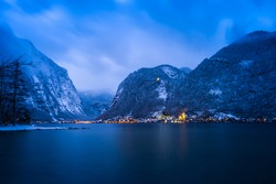 Scenery view of Austrian snowy village Hallstatt by lake surrounded by snow mountains with lights on after sunset at night time, Austria. Image contains of noise and dust from snow falling down.