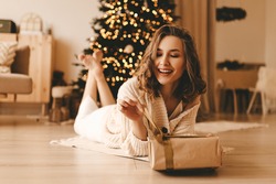 Happy emotional surprised young woman opens a holiday gift sitting on the floor by the Christmas tree in the cozy living room of the house, the concept of happiness