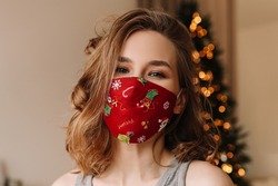 A large portrait of a smiling sick woman in a protective mask against coronavirus with a Christmas pattern stands against the background of a Christmas tree in a cozy house during the holidays