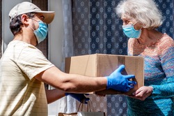 Young male volunteer in mask gives an elderly woman boxes with food near her house. Son helps a single elderly mother. Family support, caring. Quarantined, isolated. Coronavirus covid-19. Donation