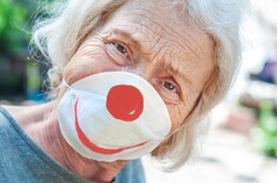 Angry sad grandmother elderly woman in a respiratory mask with a painted funny clown mask. Tired of wearing a mask. Old lady makes fun of wearing a mask, covid-19. Concept of deception coronavirus