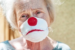 Angry grandmother elderly woman in a respiratory mask with a painted funny clown mask. Tired of wearing a mask. Old lady makes fun of wearing a mask, covid-19. Concept of deception coronavirus
