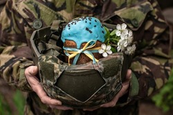 Ukrainian Easter Cake in A Hands of a Soldier in a Military Helmet. Army of Ukraine celebrate of Easter. War in Ukraine. Easter Bread with Ukraine Symbols and spring flowers. Focus on glaze