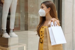 Portrait of young fashion woman wearing protective mask KN95 FFP2 with shopping bags looking through shop window