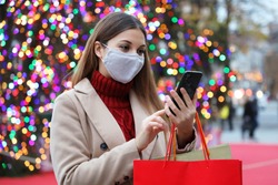 Christmas woman wearing face mask on street holding shopping bags and smart phone for online purchases with colorful christmas tree lights on background