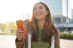 Young cheerful woman using smartphone over city background smiling looking to the side and staring away thinking