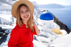 Holidays in Greece. Close-up of smiling girl in Oia, Santorini. Relaxed woman with red dress and hat looking at camera and posing in Santorini with blue dome church in the background.