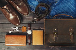 Top view, men's fashion personal belongings and accessories with space on a dark wooden background. Leather bag, shoes, watch, jeans, belt.