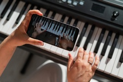 A girl watches an online music lesson on a smartphone and learns to play the piano. Close-up of woman's hands on keys.