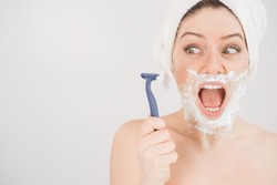 Funny portrait of a woman with shaving foam on her face holding a razor on a white background. The girl removes the mustache and beard
