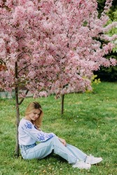 Young blonde woman rests on lawn in garden dressed in Ukrainian embroidered shirt near blooming sakura tree.
