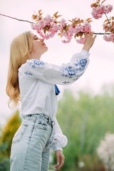 Young blonde woman sniffs flowers of blooming sakura branch in garden dressed in Ukrainian embroidered shirt and enjoys by it with closed eyes.
