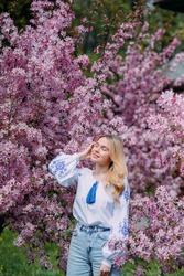 Young blonde woman walks in garden dressed in Ukrainian embroidered shirt and enjoys by blooming sakura trees with closed eyes.