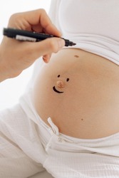 Couple parents has fun and draws smiley with felt pen on pregnant belly of woman. Concept of love and care during pregnancy, expecting and born baby.