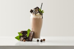 Chocolate milkshake with cream decorated chocolate portions and balls around on white wooden table and light isolated background. Front view.