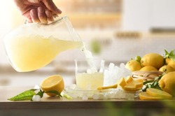 Person pouring glass with fresh lemon drink with ice from pitcher on kitchen table with lemons and crushed ice. Front view.