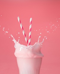 striped straws in a glass of splashing strawberry milkshake isolated on pastel pink color backdrop