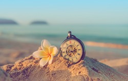 retro pocket watch and plumeria flower on sand sea beach, tropical ocean background. summer vacation, relax time, travel concept. close up