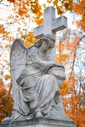 Angel statue with cross on old cemetery, autumn natural background. Design for condolences, mourning cards or obituary. concept of religion, faith in God, mourn, memory. all saints day, All Souls Day