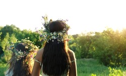 Two girls in flower wreaths on meadow, sunny green natural background. Floral crown, symbol of summer solstice. Slavic ceremony on Midsummer, wiccan Litha sabbat. pagan holiday Ivan Kupala