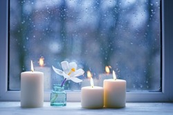 Burning candles, white flower in bottle on windowsill, abstract blurred raindrops glass background. spring season. Melancholy cozy mood. Relax, harmony, meditation, life balance concept. rainy weather